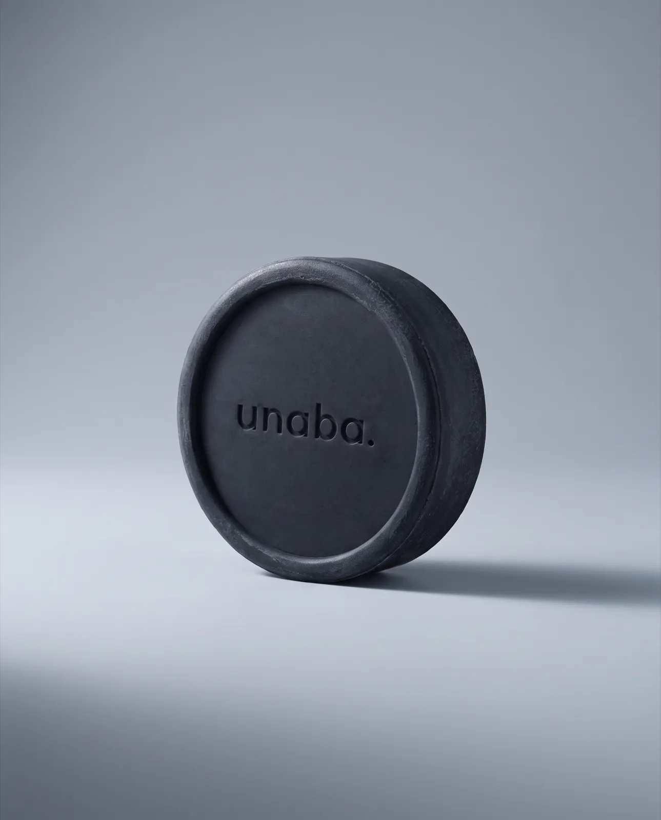 unaba Miracle Shampoo Bar — matte black puck, 15° front view on a cool-grey studio sweep